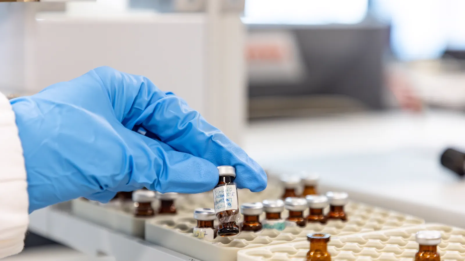 Gloved hand selecting an amber sample vial from a tray in a research laboratory, illustrating BPC-157 research
