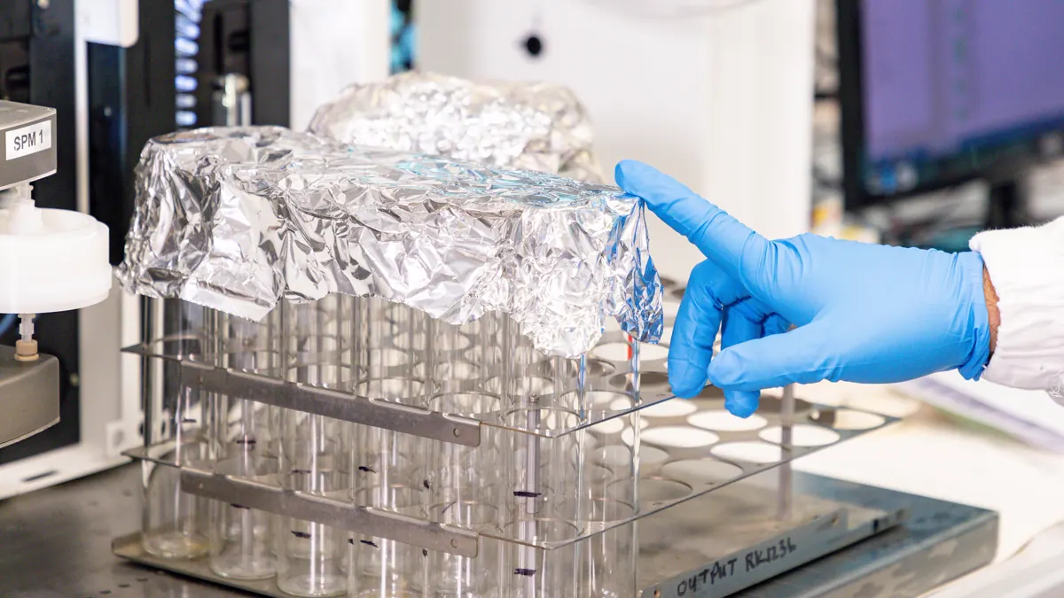 Gloved hand lifting foil from a rack of glass sample tubes during side-by-side laboratory preparation